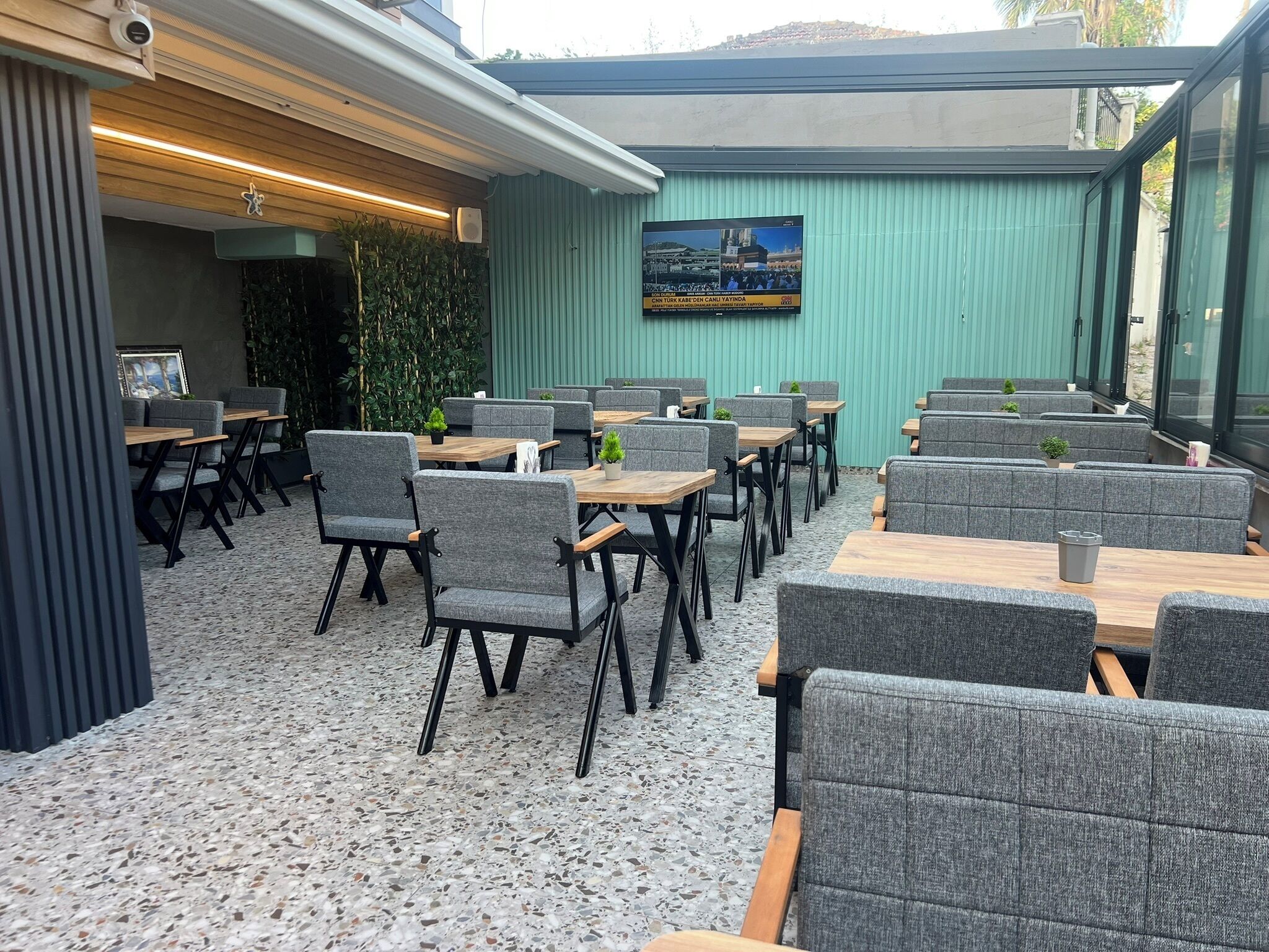 An outdoor seating area with tables and chairs, a television screen, and a green wall.