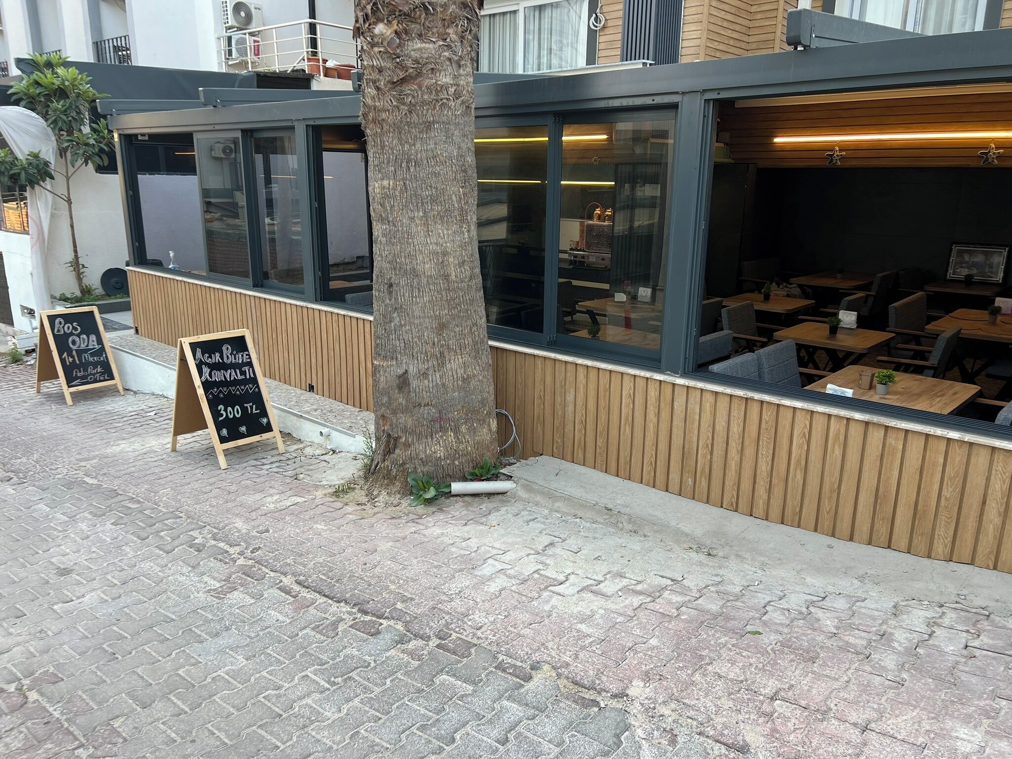 Un área de estar al aire libre con mesas y sillas de madera, un árbol y dos pizarras con texto.
