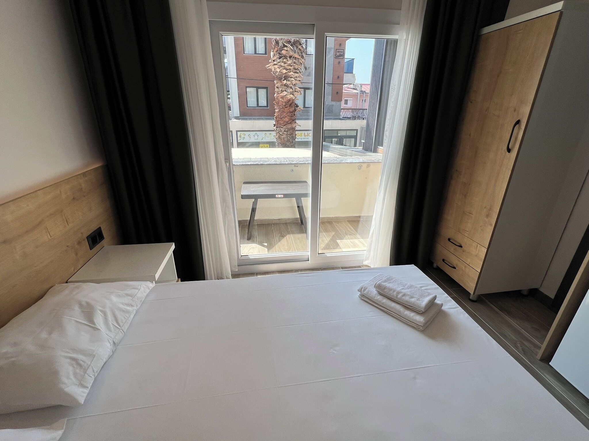 A hotel room with a bed, a wooden headboard, a wardrobe, and a sliding glass door leading to an outdoor balcony.
