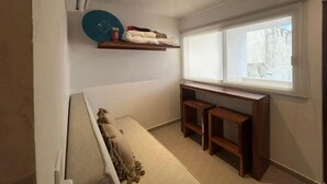 Une petite pièce bien éclairée, avec une fenêtre, un lit, une étagère où sont pliés des vêtements et un bureau avec deux chaises.