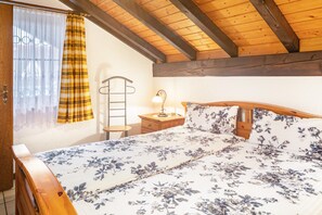 Quarto com teto de madeira, cama com roupa de cama floral, mesa de cabeceira com abajur e janela com cortinas.