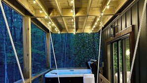 En træterrasse med seng og jacuzzi, omgivet af glasvægge med udsigt til skov.