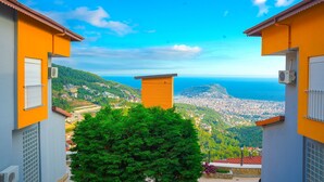A view of a coastal city with a clear sky, featuring buildings with orange and grey facades, a prominent tree in the foreground, and a distant view of the sea and cityscape.