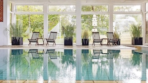 Une piscine intérieure aux parois en verre transparent, entourée de plantes en pot et de fauteuils en osier.