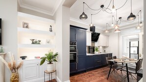 A modern kitchen with dark blue cabinets, white countertops, and a herringbone wood floor. There are pendant lights, a dining table with chairs, and a potted plant. - GuestReady – Stylish 3bed in Central Pimlico (London)