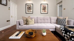 A living room with a light grey sectional sofa, two framed artwork pieces, a wooden coffee table with a magazine, remote controls, a woven basket, and a potted plant. - GuestReady – Stylish 3bed in Central Pimlico (London)