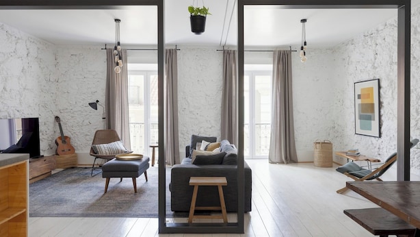 Ein modernes Wohnzimmer mit einem grauen Sofa, einem Holztisch, einem Korbstuhl, einem kleinen Hocker und einer Gitarre. Ein Fernseher, eine hängende Pflanze und ein gerahmtes Bild sind an der Wand zu sehen.