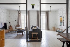 A modern living room with a grey sofa, a wooden coffee table, a wicker chair, a small stool, and a guitar. There is a television, a hanging plant, and a framed picture on the wall. - Architectural Gem in Bica II (Lisboa)