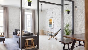 Ein modernes Wohnzimmer mit einem grauen Sofa, einem Holzschemel und einem Esstisch aus Holz. Es gibt zwei hängende Pflanzen, ein gerahmtes abstraktes Gemälde und einen gewebten Korb auf dem Boden.