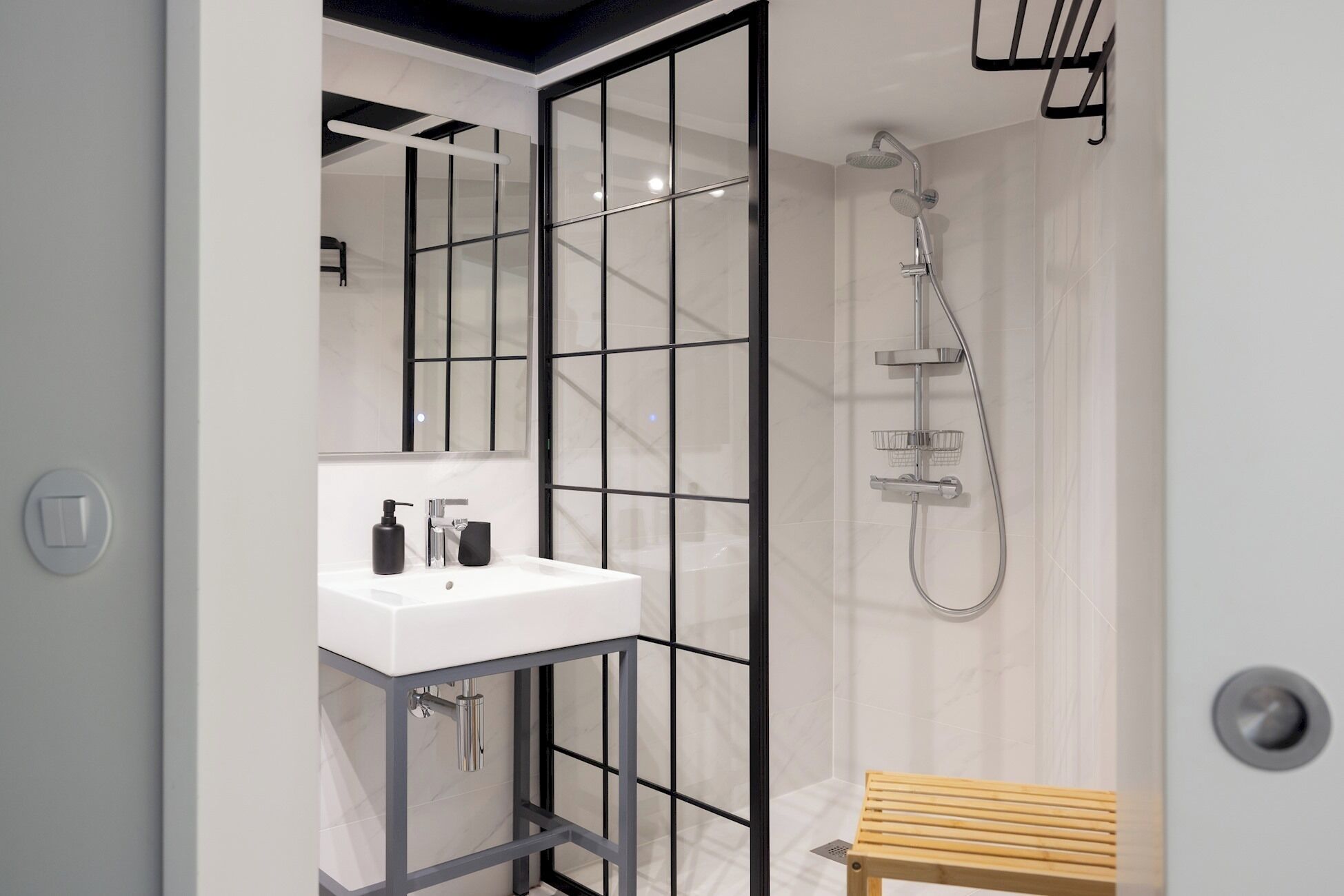 A modern bathroom with a white sink, black fixtures, a glass shower enclosure, and a wooden bench.