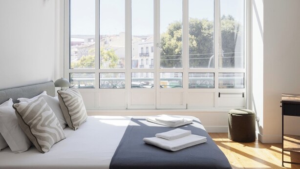 A bedroom with a large window, a bed with pillows, a bedside table, and a trash bin.