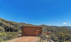 Une cabane en bois, accessible par un sentier de terre, entourée d’une végétation luxuriante et avec des montagnes en arrière-plan.