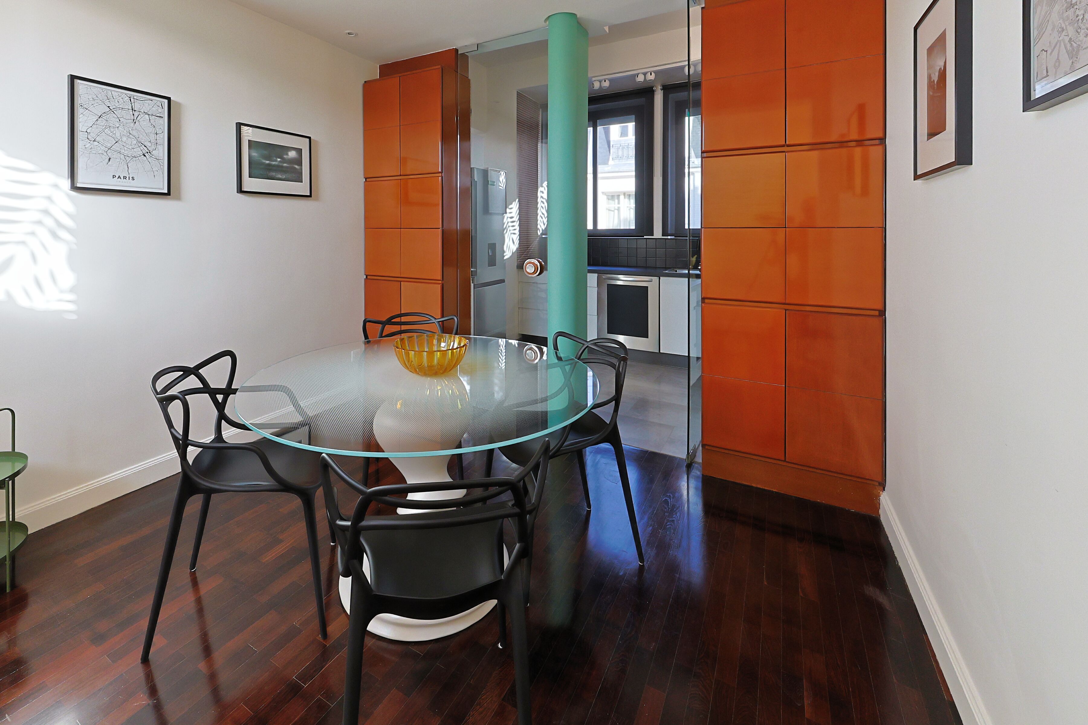 Een eethoek met een glazen tafel en stoelen, een blauwgroene zuil en een keuken op de achtergrond.