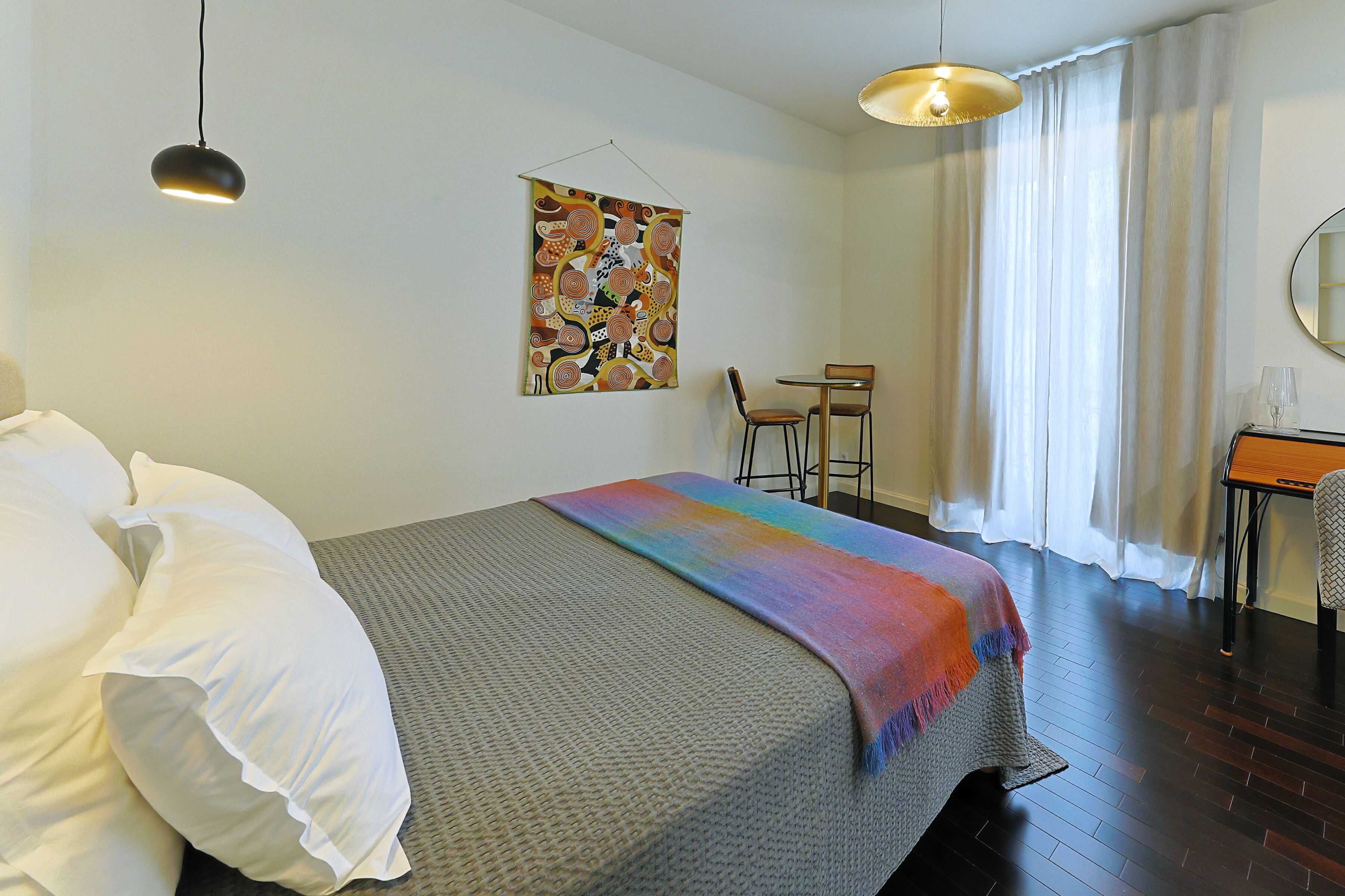 Une chambre d’hôtel avec un lit, un bureau, une chaise, une lampe suspendue et une fenêtre avec des rideaux.