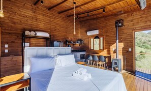 A wooden cabin with a bed, a small kitchenette, and a view of the outdoors. - Cabana aconchegante c/ jacuzzi e lareira – VST019 (Anitapolis)