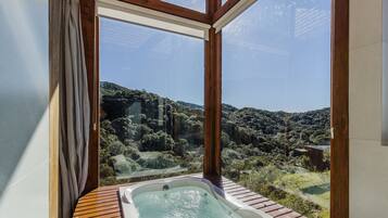 A hot tub with a wooden deck, a window with curtains, and a view of a forested hillside.
