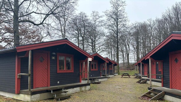Uma fileira de cabanas com detalhes vermelhos e portas numeradas, situada numa área arborizada com mesas de piquenique e uma grande rocha em primeiro plano.