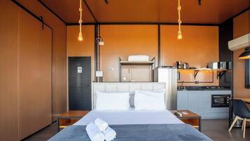A modern hotel room with a bed, bedside tables, a kitchenette, and a wooden ceiling.