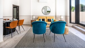 Una habitación de hotel moderna con sofá, dos sillones, una mesa de comedor con sillas, un espejo redondo y un ventanal grande.