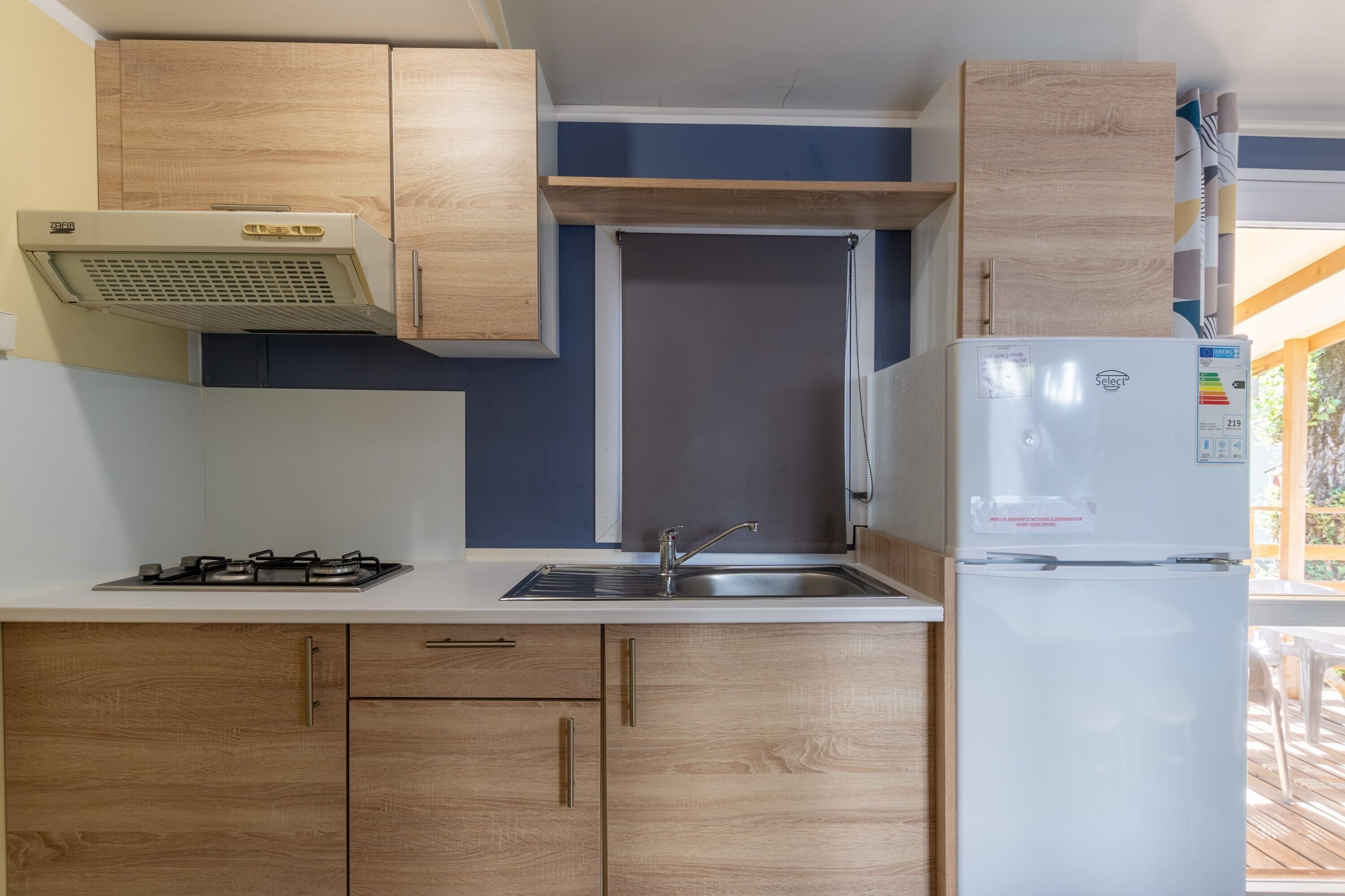 Een kleine keuken met houten kastjes, een gasfornuis, een spoelbak en een koelkast.