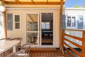Un espace extérieur abrité doté d’une terrasse en bois, d’une table avec des chaises et d’une porte-fenêtre coulissante donnant sur un espace intérieur avec une table bleue et des tabourets.