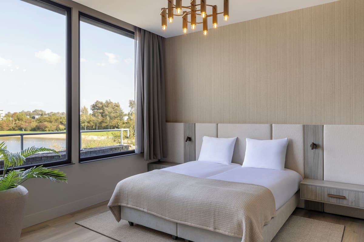 A modern bedroom with a large bed, a nightstand, a bedside table, a potted plant, and a view of a lake.