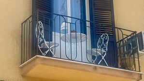 Un balcón con una mesa y dos sillas, una ventana con contraventanas y una barandilla de metal.