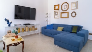 Una sala de estar con un sofá azul, un televisor, una mesa de centro de vidrio y objetos decorativos en un estante.