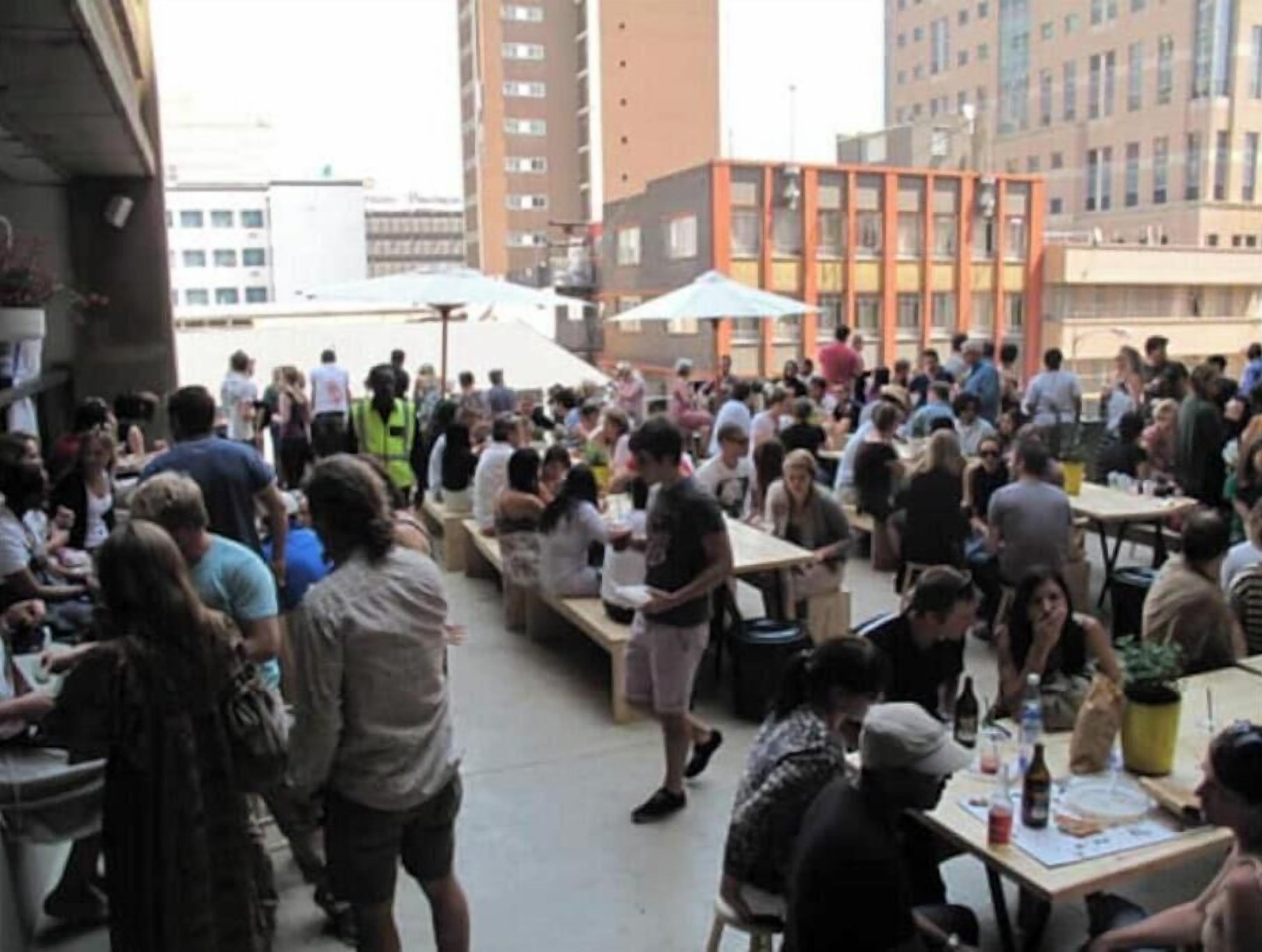 An outdoor dining area with multiple tables and chairs, surrounded by buildings.