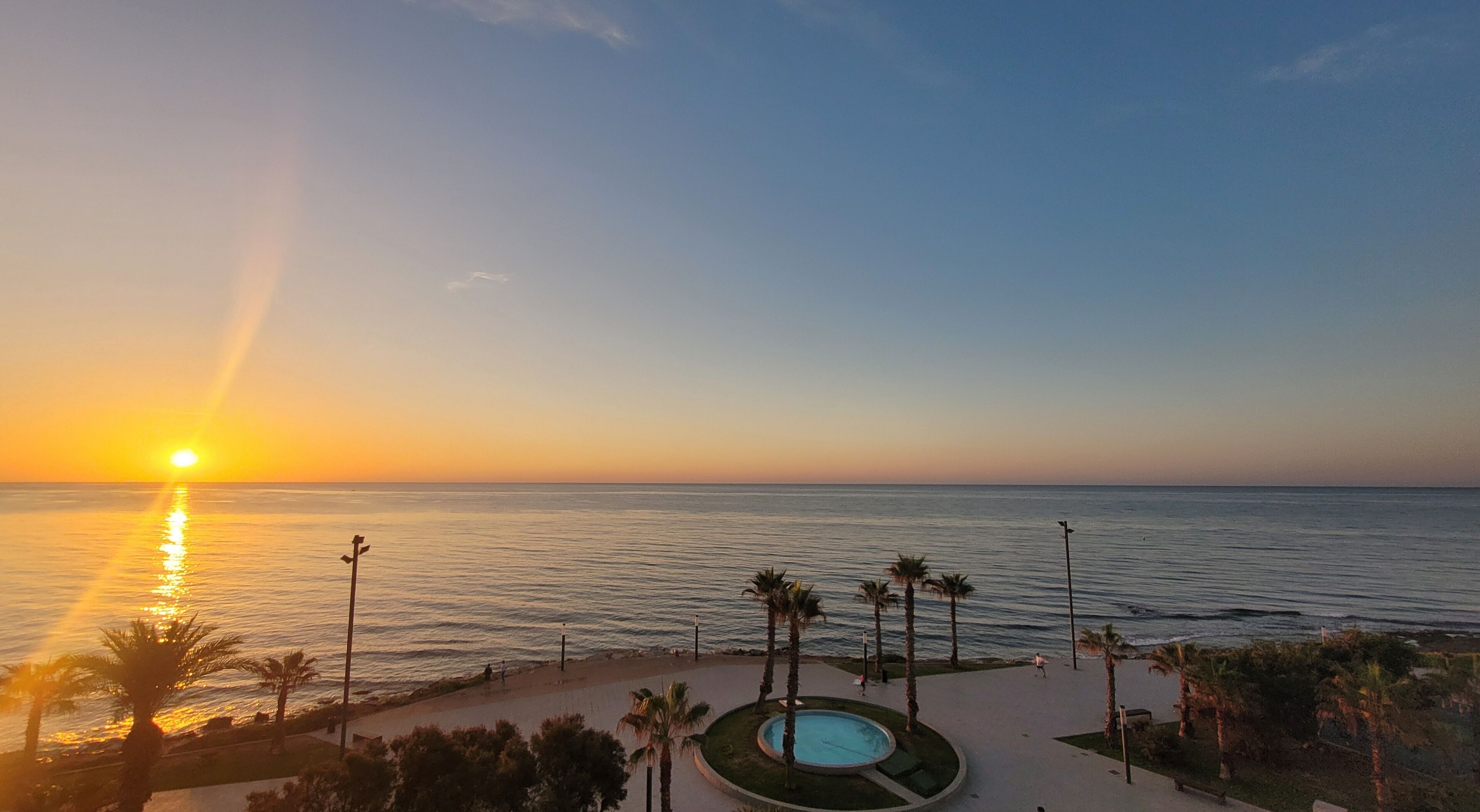 Tramonto su un mare calmo, con una piscina e palme sullo sfondo.