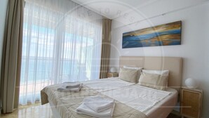Habitación de hotel con una cama grande, una mesita de noche, un cuadro y vistas al mar a través de cortinas translúcidas.