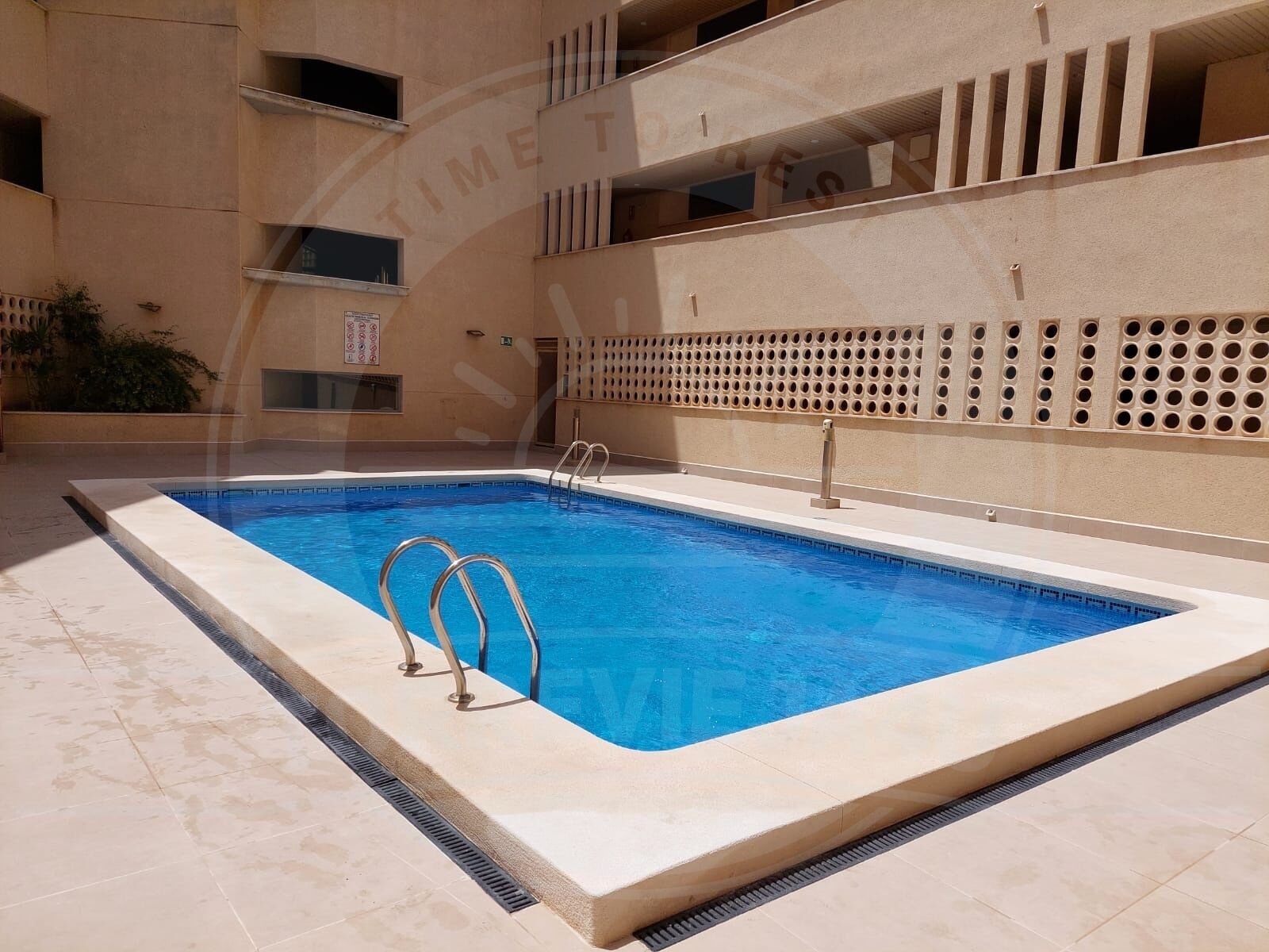 A rectangular swimming pool with a tiled deck, surrounded by a beige building with balconies and a perforated wall design.