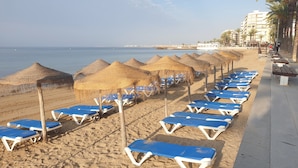 Una playa con hileras de sillas azules y sombrillas de paja, una orilla arenosa y un mar tranquilo al fondo.