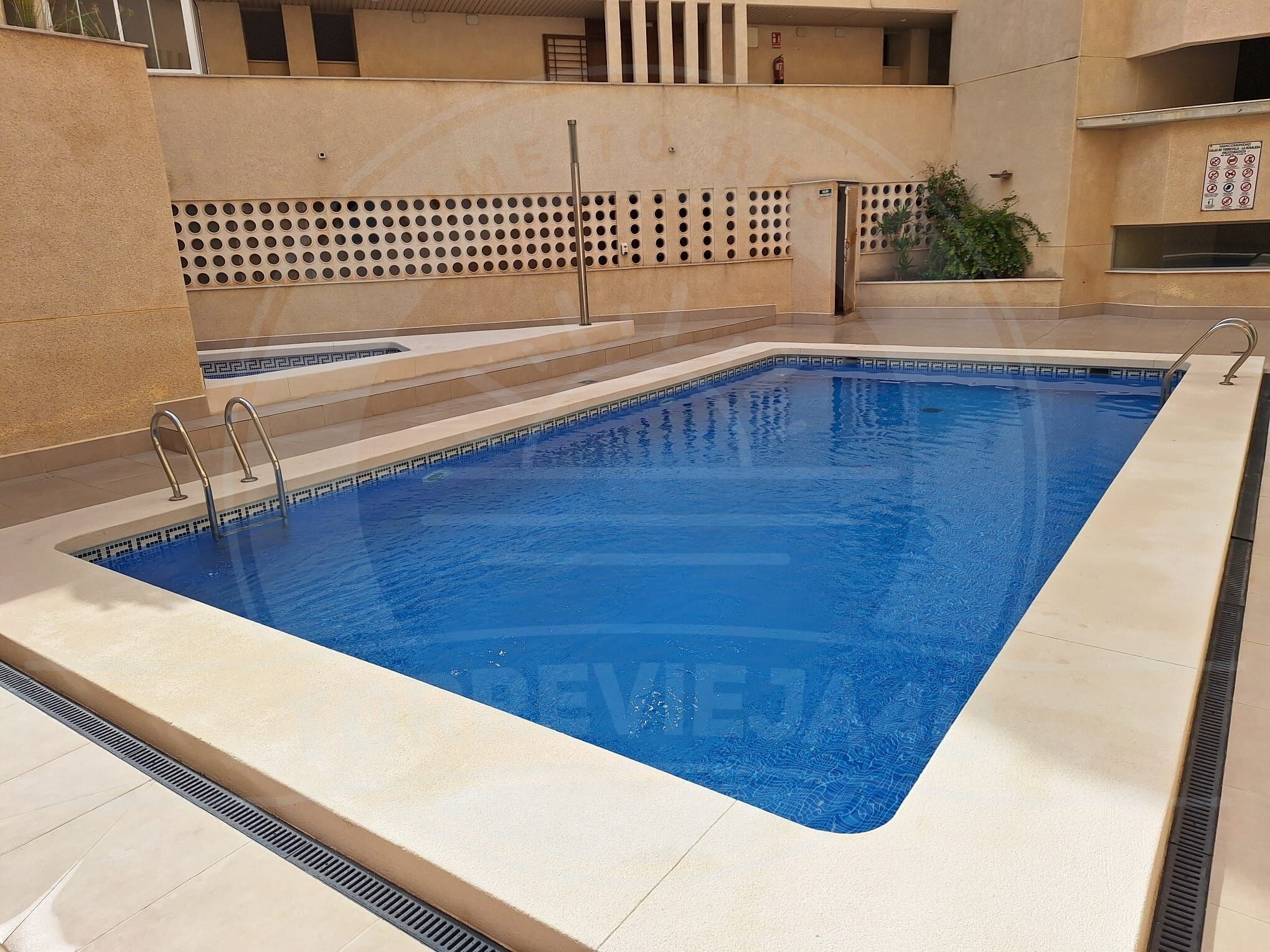 Una piscina rectangular con un bordeado de baldosas, barandales metálicos y una pared perforada al fondo.