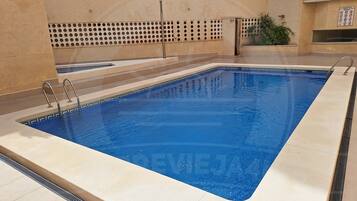 Una piscina rectangular con un bordeado de baldosas, barandales metálicos y una pared perforada al fondo.