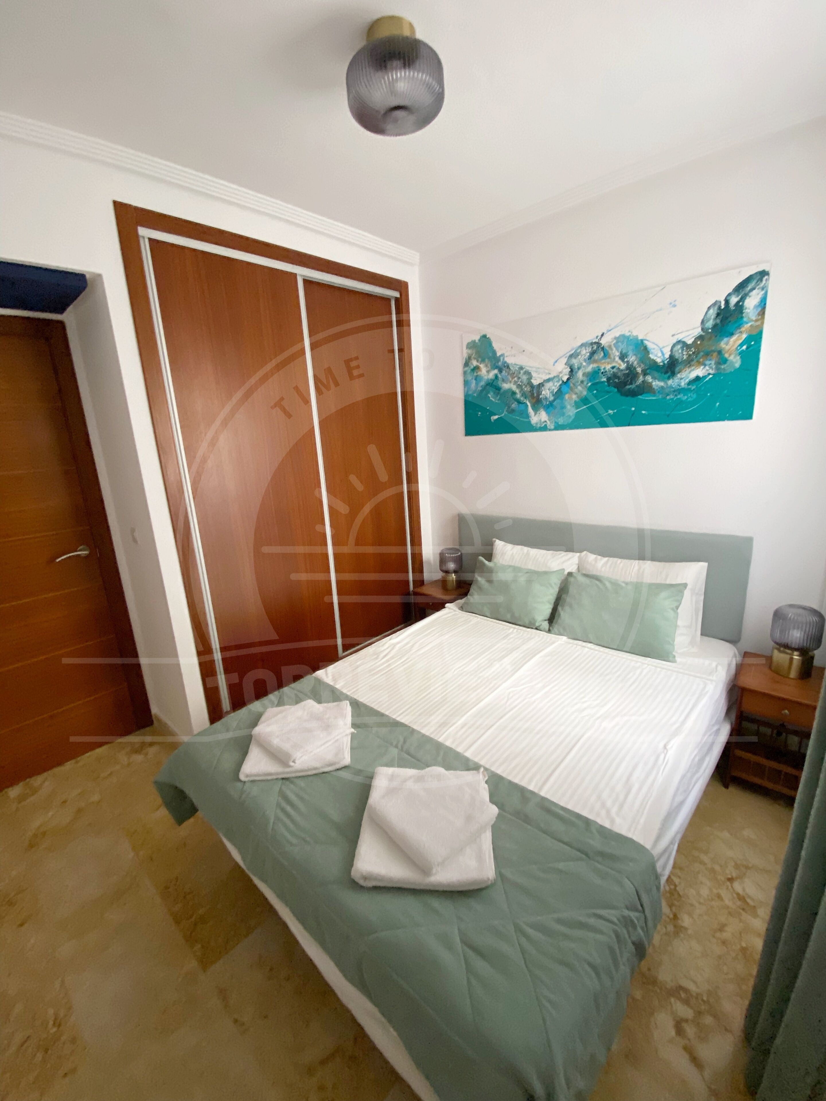 Una cama bien hecha con una colcha verde y almohadas blancas, un armario de madera, una mesita de noche con una lámpara y un cuadro en la pared.