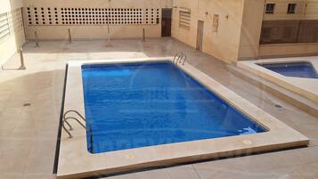 Una piscina rectangular con una zona de terraza empavedada, una escalera metálica y un pequeño jacuzzi adyacente a la piscina.