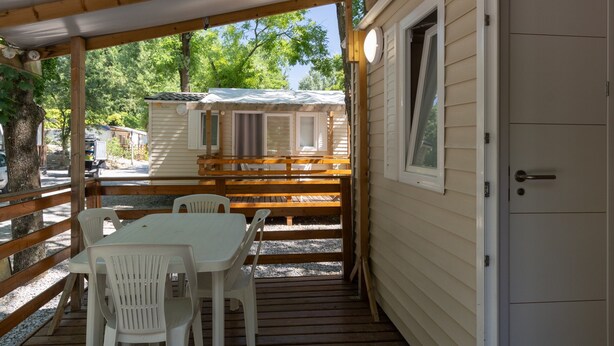 A covered outdoor area with a table and chairs, a wooden deck, and a building with a door. - Mobile Home 'Mobil-home D - 1-4 Pers' with Shared Pool and Shared Garden (Lavilledieu)