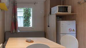 A compact room with a wooden table, a white chair, a microwave, a refrigerator, and a window with curtains. - Mobile Home 'Mobil-home D - 1-4 Pers' with Shared Pool and Shared Garden (Lavilledieu)