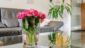 A modern living room with a glass coffee table, a grey sofa, and a vase of pink roses. - Modern Apartment in Dream Location with Mountain View, Shared Garden and Wi-Fi (Langelsheim)