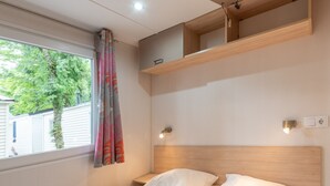 Une chambre à coucher comprenant un lit, une table de chevet, une étagère fixée au mur, une fenêtre avec des rideaux et une vue sur un arbre à l’extérieur.