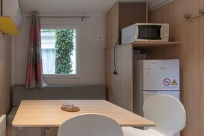 Ein gemütlicher Raum mit Holztisch, weißem Stuhl, einer Mikrowelle, einem Kühlschrank und einem Fenster mit Vorhängen.