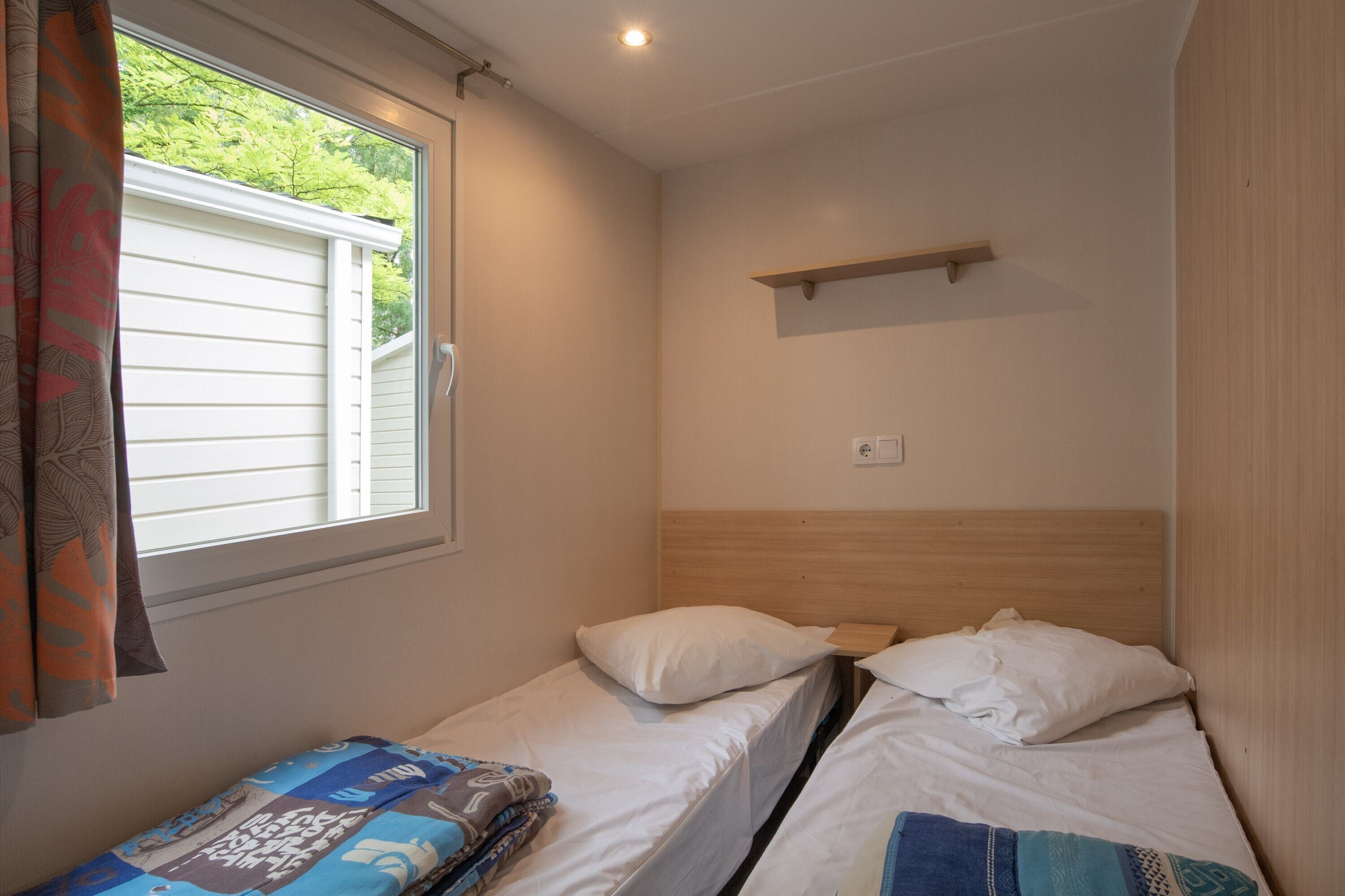 A small, single-bed room with a window, a wooden headboard, and a wall-mounted shelf.