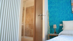 Une chambre d’hôtel avec une douche, un lit recouvert d’une couvre-lit à motifs bleus et blancs, une table de chevet avec une lampe, et un mur orné d’un motif texturé bleu.