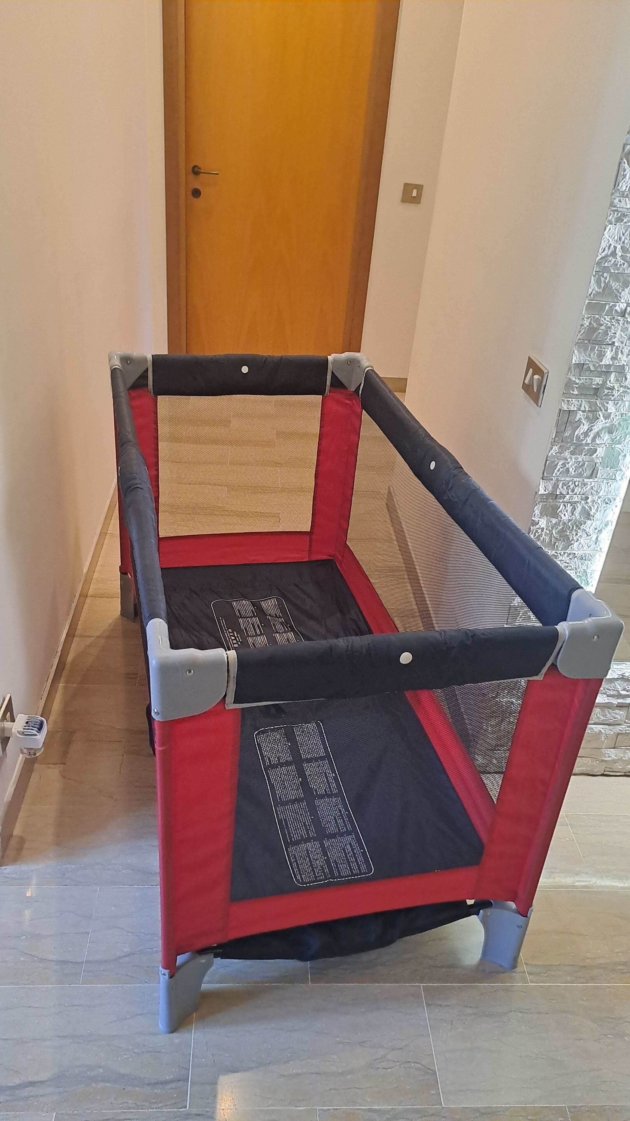 Ein kleiner, abgegrenzter Spielbereich mit rotem und schwarzem gepolsterten Boden, grauen und beigen gepolsterten Wänden und einer Holztür im Hintergrund.