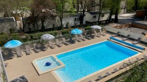 Una piscina rectangular con un jacuzzi pequeño, rodeada de sillas de descanso y sombrillas, con árboles y edificios al fondo.
