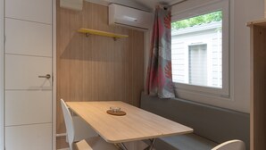 Una habitación pequeña con una mesa de madera, un banco, una silla blanca, un equipo de aire acondicionado y una ventana con cortina.
