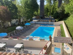 A rooftop swimming pool with lounge chairs and umbrellas, surrounded by greenery and a building with a chimney.