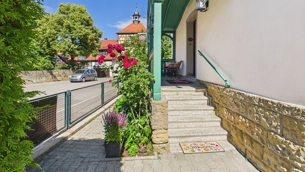 Un escalier mène à un bâtiment doté d’une marquise verte et d’une lanterne. Des plantes en pot, ornées de fleurs, se trouvent au pied de l’escalier.