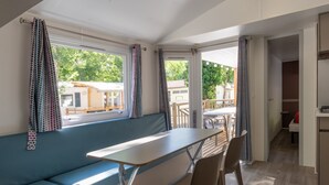 A room with a bench, table, and chairs, large windows with curtains, and an open door leading to another area. - Mobile Home 'Confort A - 1-8 Pers' with Shared Pool, Private Terrace and Shared Garden (Lavilledieu)